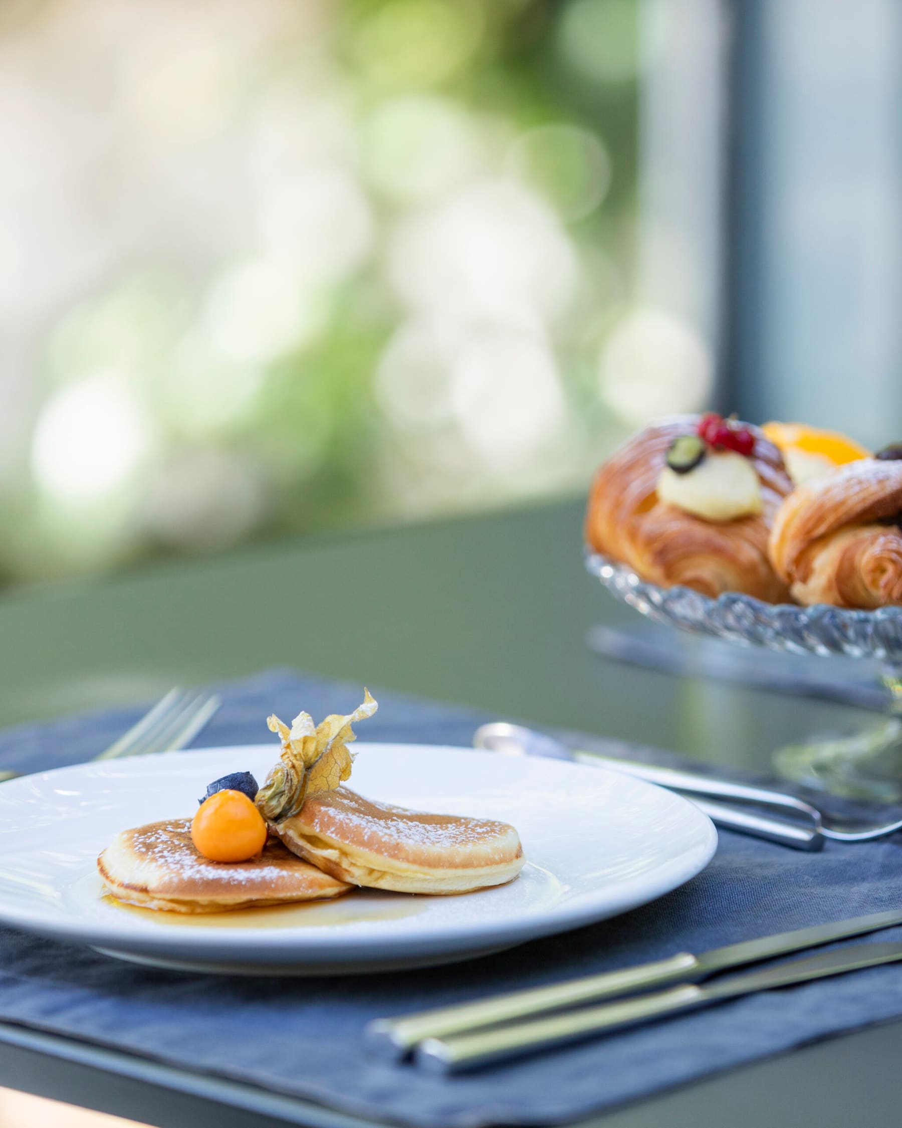 Pancake e cornetti su un tavolo verde salvia, con lino grigio azzurro e alzata di vetro