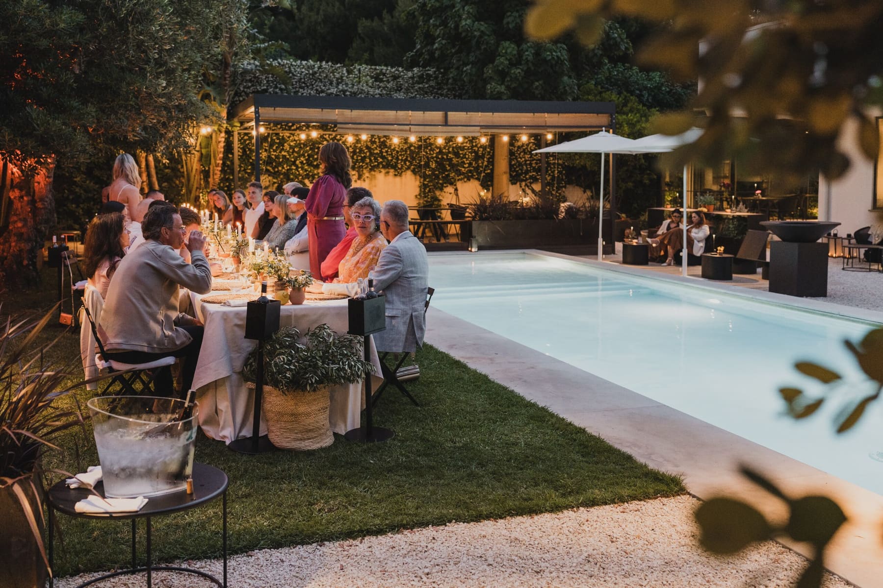 La cena servita in giardino dopo il tramonto: tavoli apparecchiati a bordo piscina, lucine appese fra gli alberi e ospiti seduti a tavola